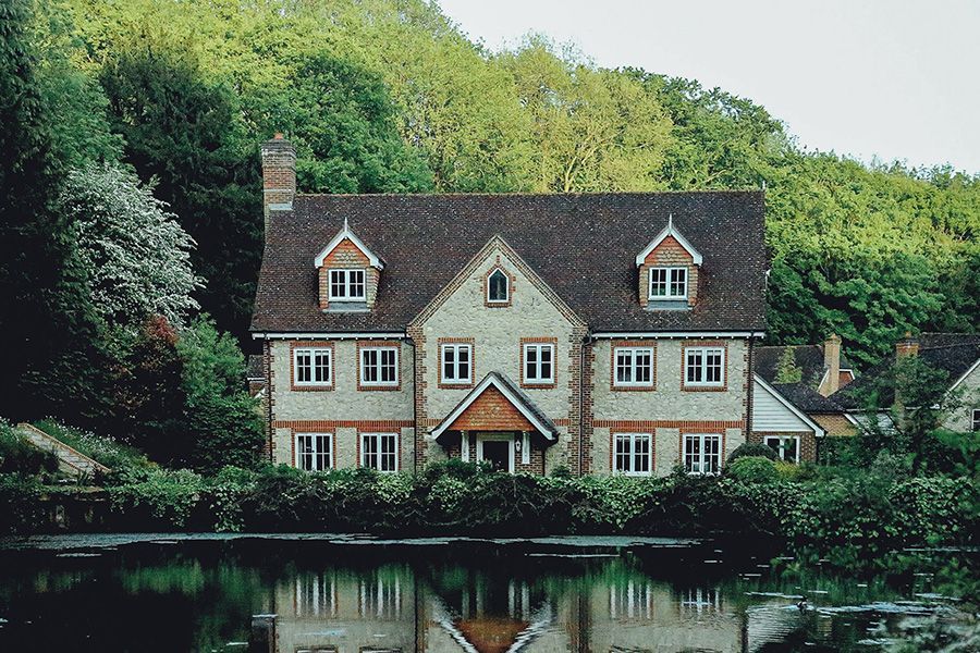 Cottage-style house with a red roof beside a calm lake, reflected in the water and surrounded by trees