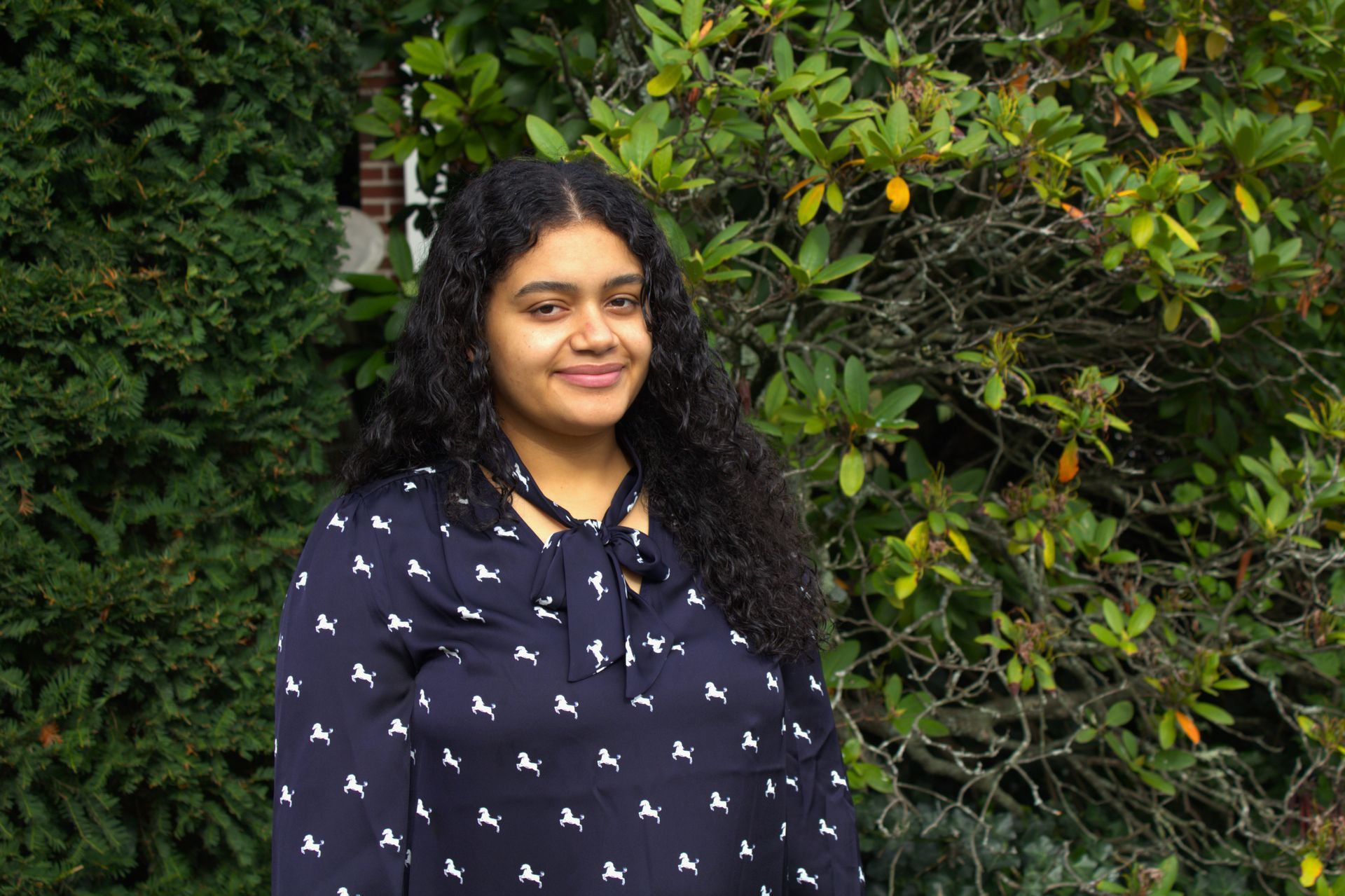 A woman in a blue shirt is standing in front of a bush.