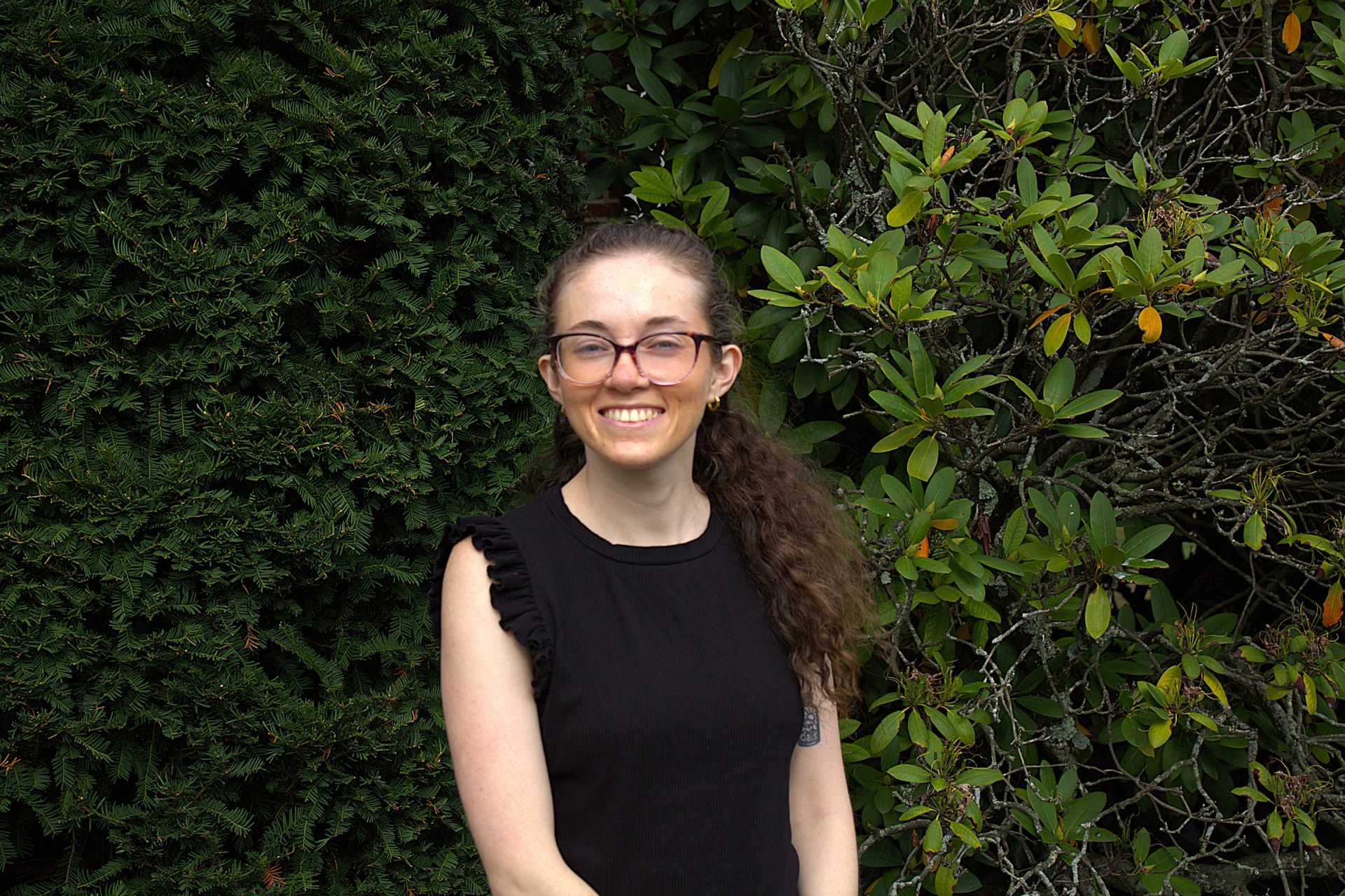 A woman wearing glasses and a black shirt is standing in front of a bush.
