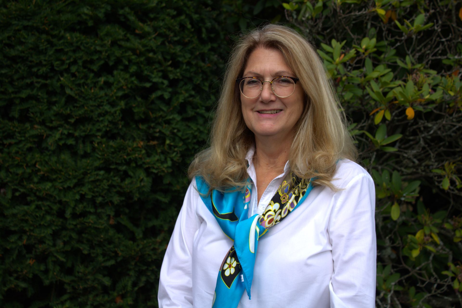 A woman wearing glasses and a blue scarf is standing in front of a bush.