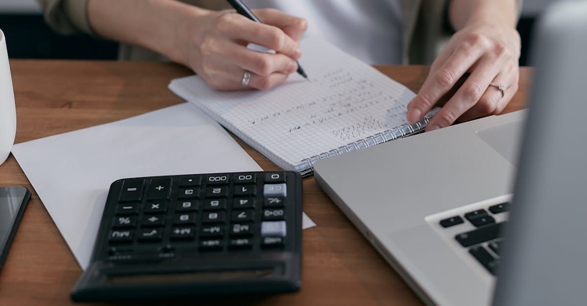 A woman is writing in a notebook next to a laptop and a calculator.