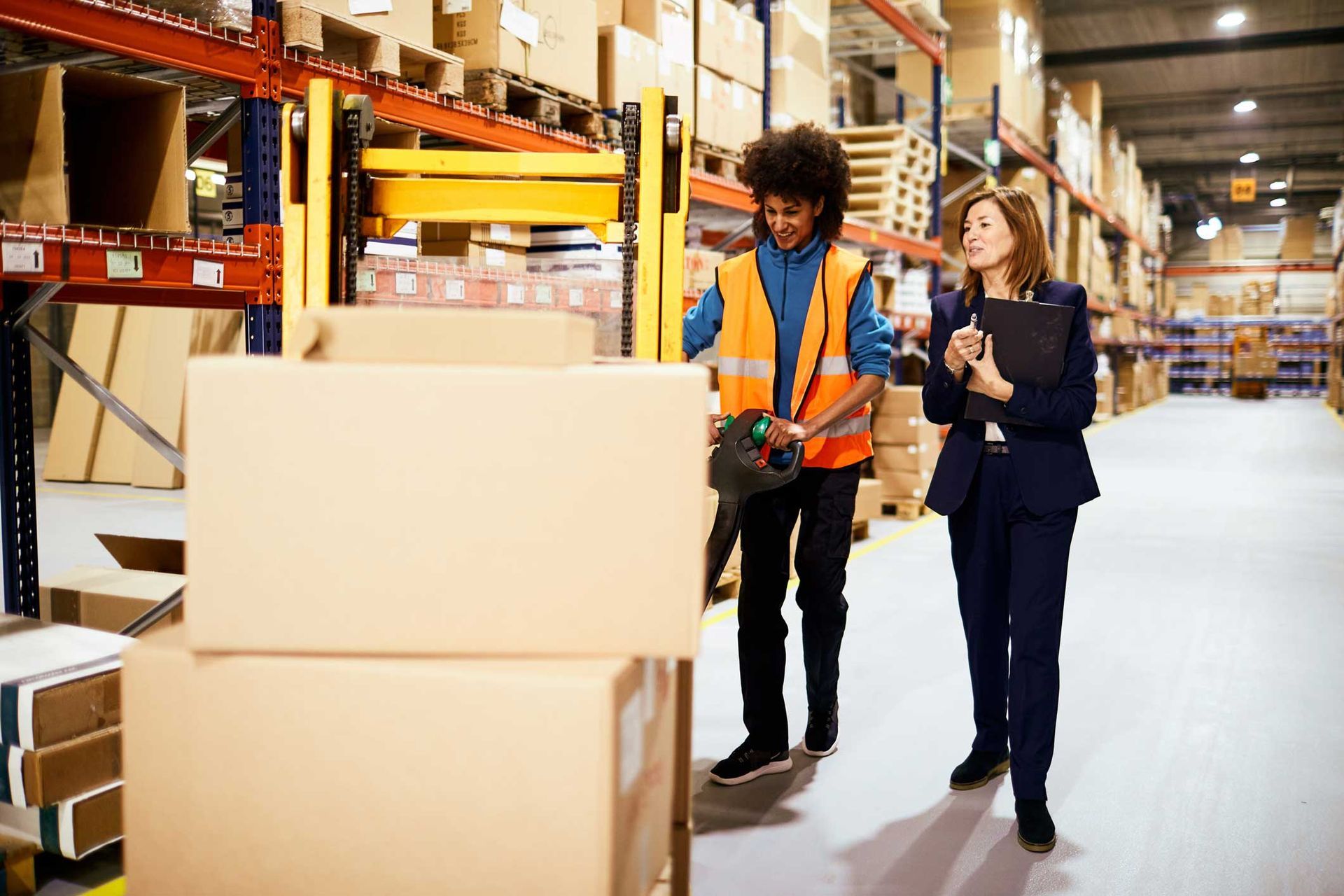 Warehouse worker in orange vest and businesswoman with clipboard walking through aisles of stacked boxes. Warehouse worker in orange vest and businesswoman with clipboard walking through aisles of stacked boxes.