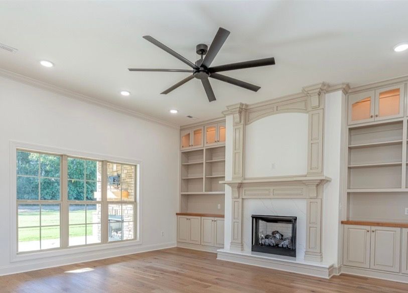 Bright living room with built-in shelves, fireplace, large window, wood floors, and a black ceiling fan