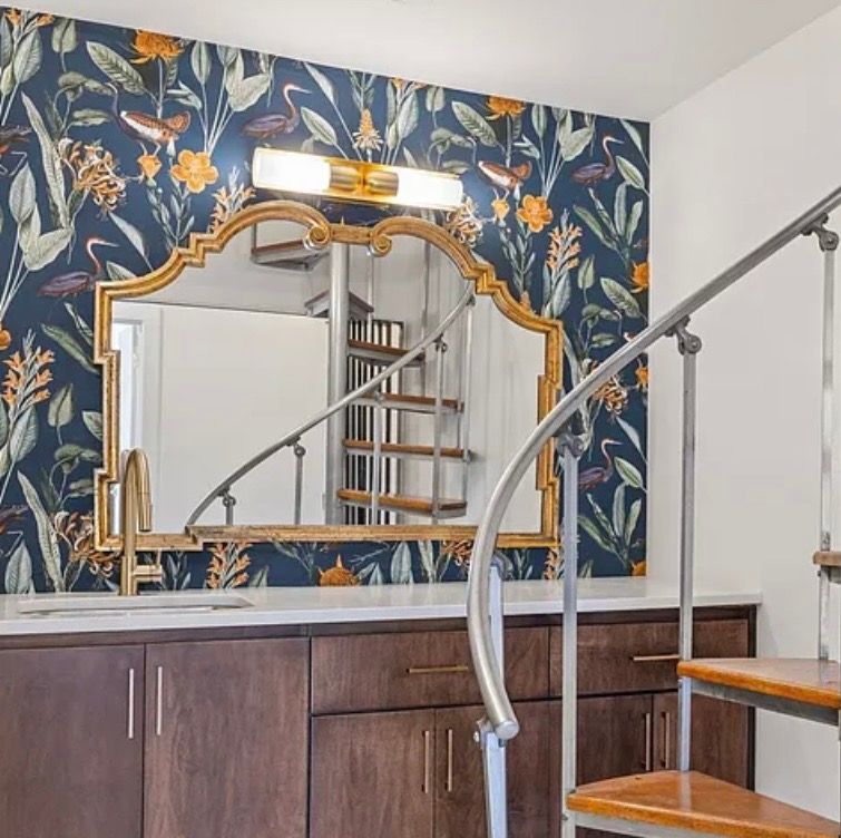 Stylish stair landing with floral wallpaper, ornate gold mirror, and wood railing under a wall light