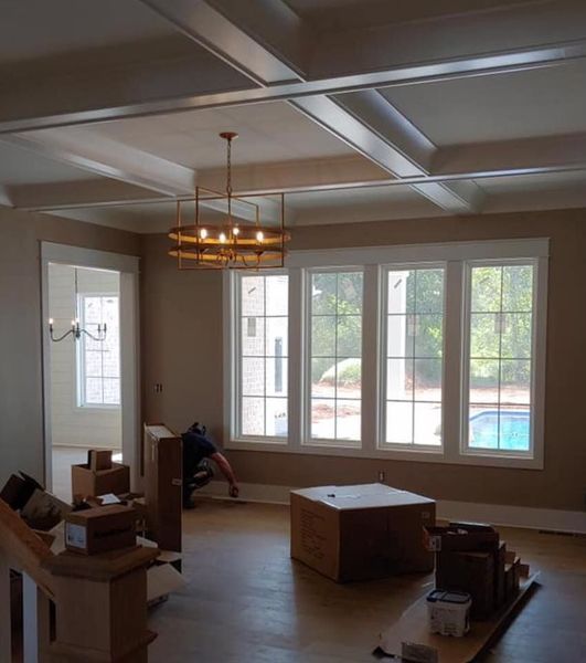 Spacious living room with large windows, a coffered ceiling, and boxes stacked on the floor during a move.