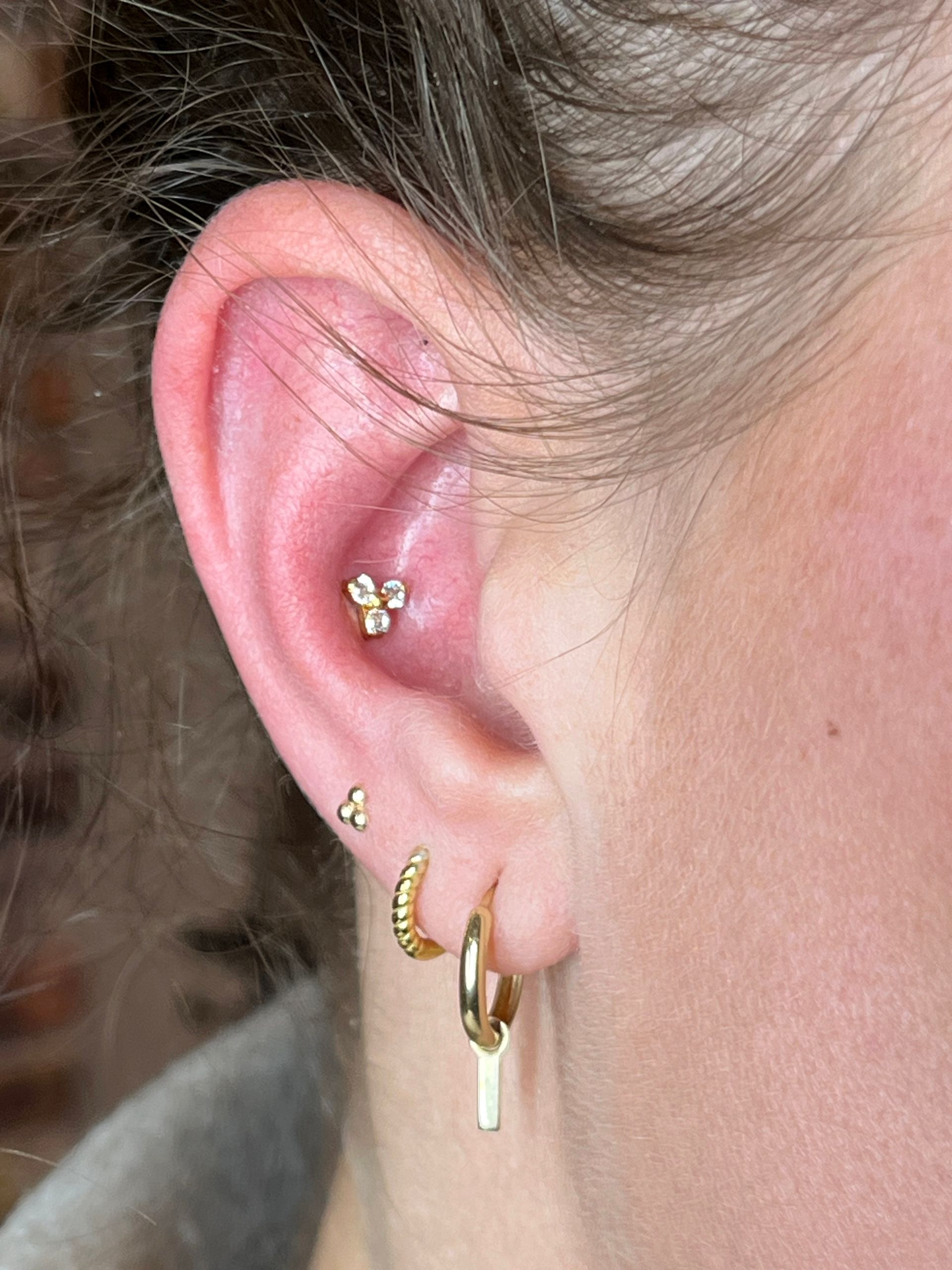 Een close-up van iemands oor met een gouden oorsteker en drie gouden oorbellen in de oorlel.