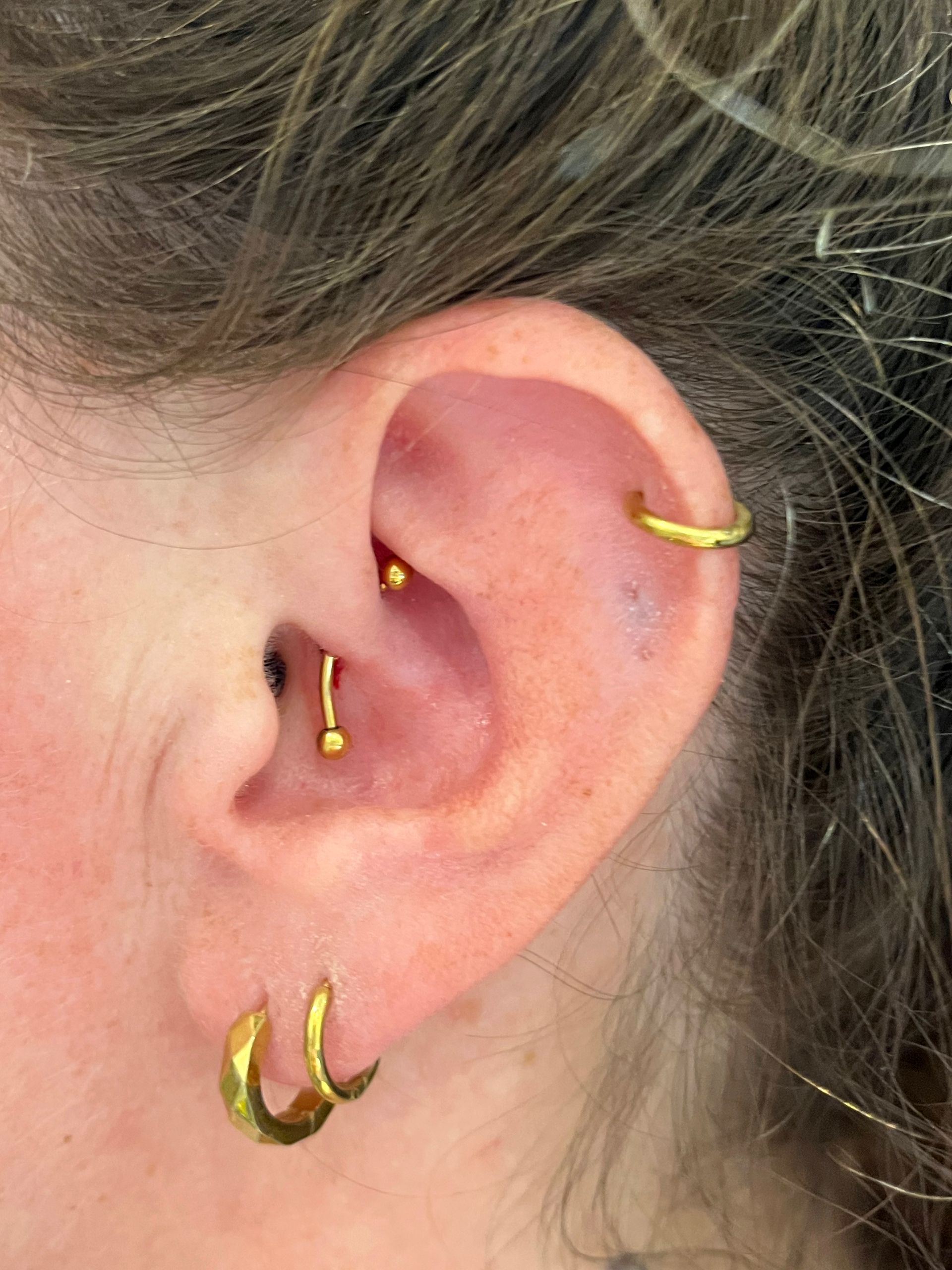 Een oor versierd met gouden sieraden, waaronder twee oorringetjes, een helixringetje en een rookpiercing met een gebogen staafje.