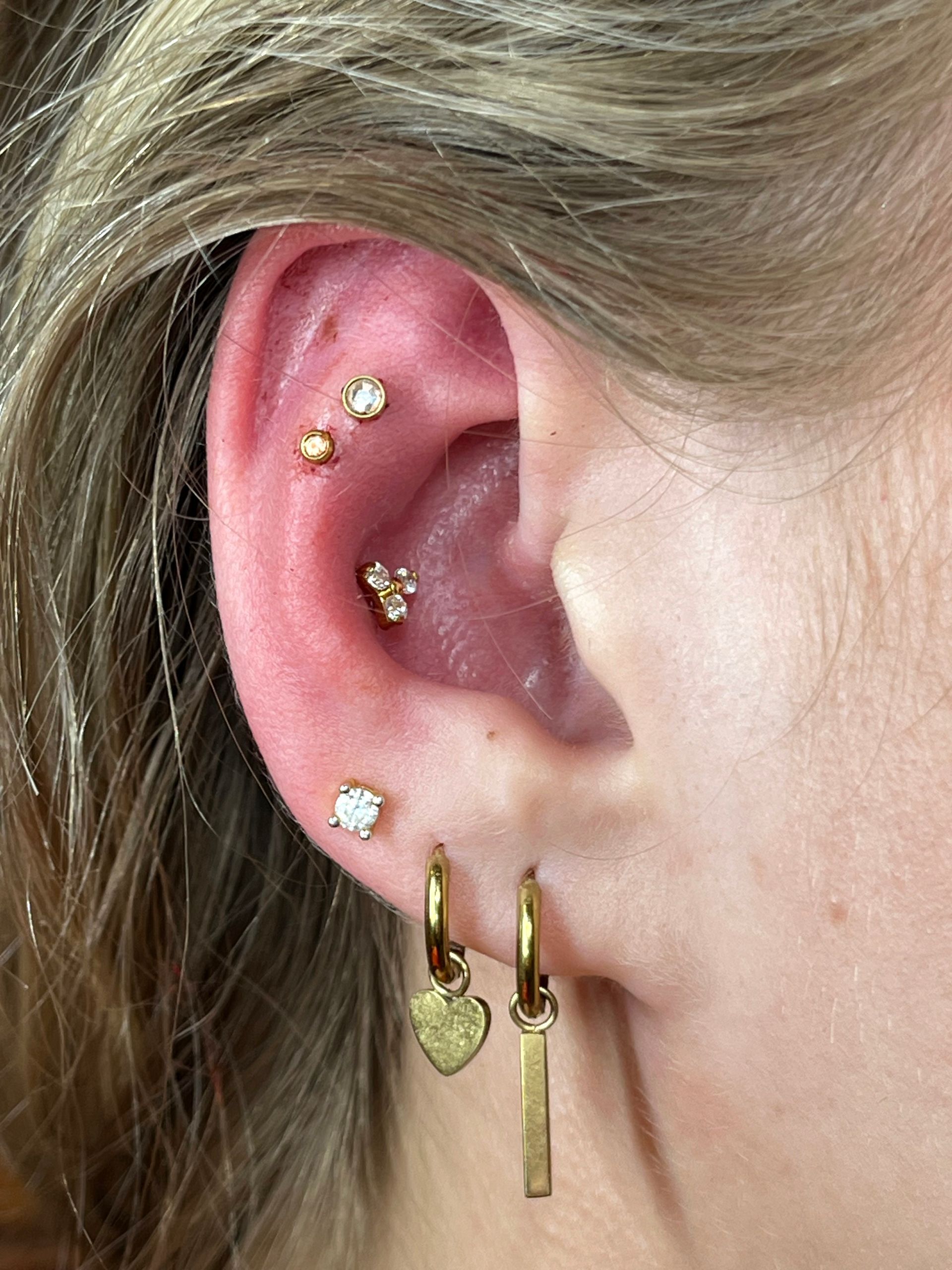 Een close-up van een oor met meerdere gouden piercings, waaronder een conch-piercing, twee platte piercings, een oorlel-piercing en twee hangende oorringen.