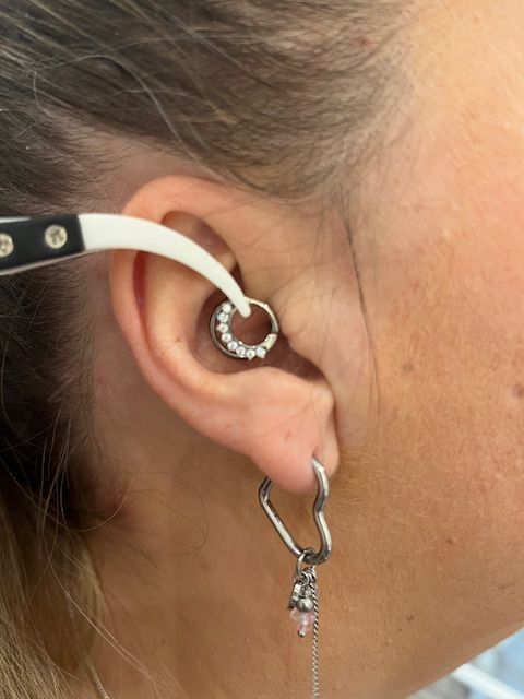 Een instrument houdt een decoratieve ringpiercing in iemands oor vast. Daaronder hangt een hartvormige oorbel aan de oorlel.