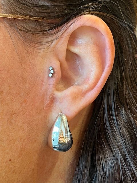 Een close-up van een oor met een zilveren traguspiercing, drie kleine, op elkaar gestapelde kraaltjes en een grote, druppelvormige oorbel.