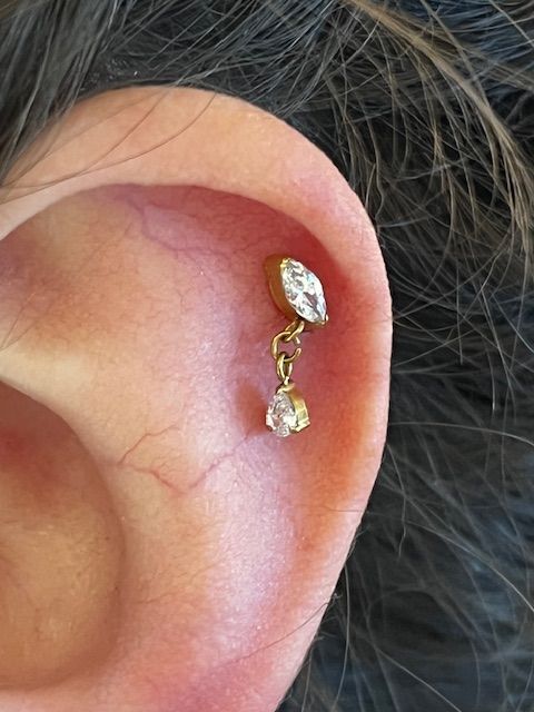 Een close-up van menselijk oorkraakbeen met een gouden, marquisevormige oorbel met een klein, hangend peervormig steentje.