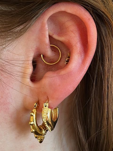 Een close-up van een oor met een gouden daith-ring, een klein kristallen oorknopje en twee goudkleurige oorringen met textuur.