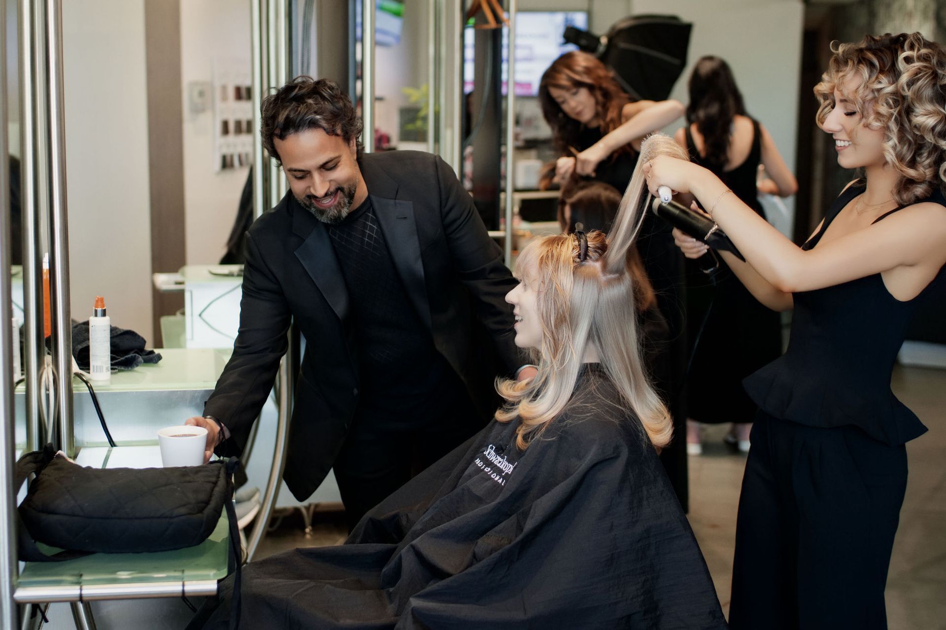 Hair stylists work on a client in a salon. One is blow-drying, others are adjusting hair.