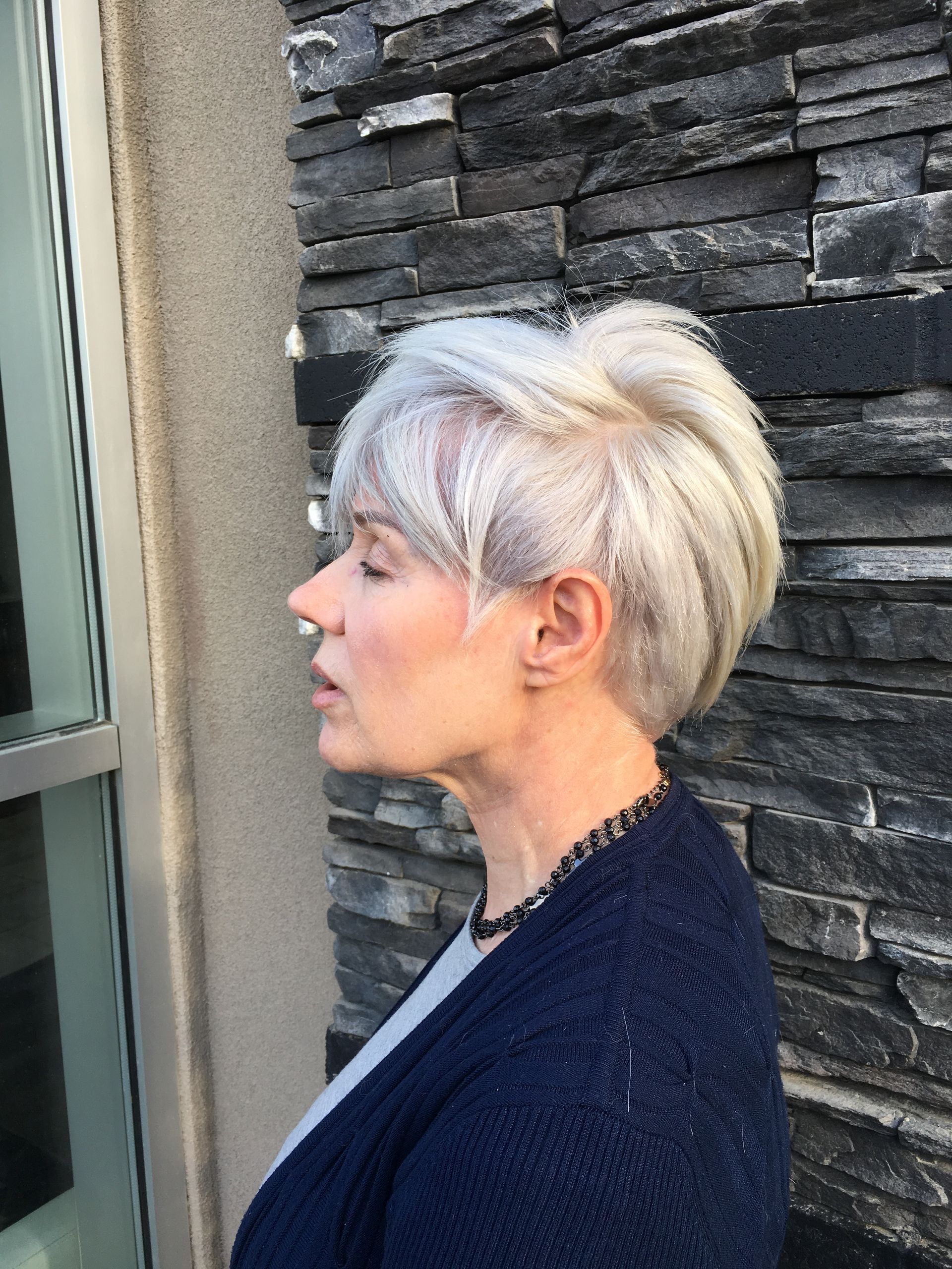 A woman with short white hair is standing in front of a stone wall.
