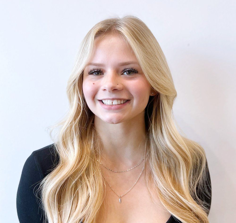 A woman with blonde hair is wearing a black shirt and a necklace and smiling.