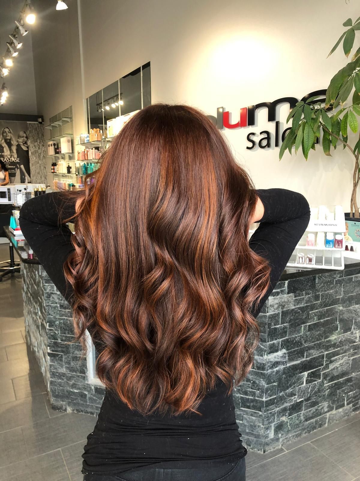 A woman with long brown hair is standing in a salon with her hands behind her head.