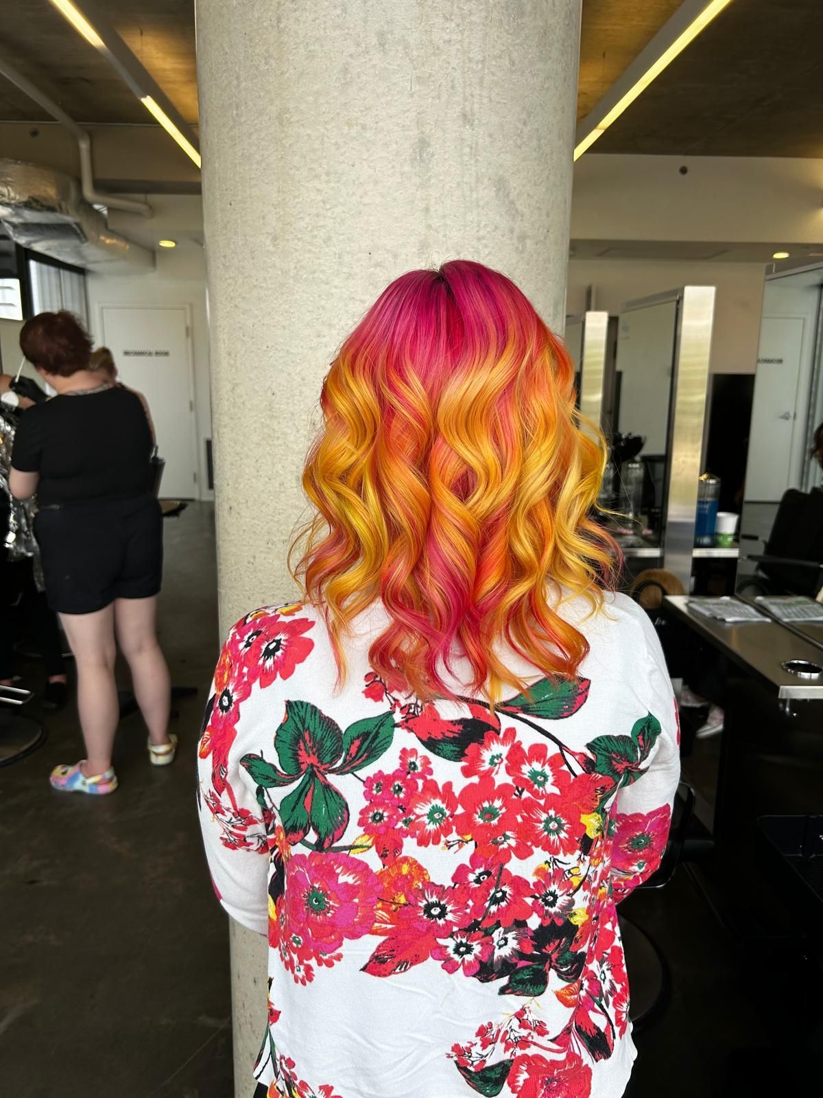 A woman with brightly colored hair is standing in front of a pillar in a salon.
