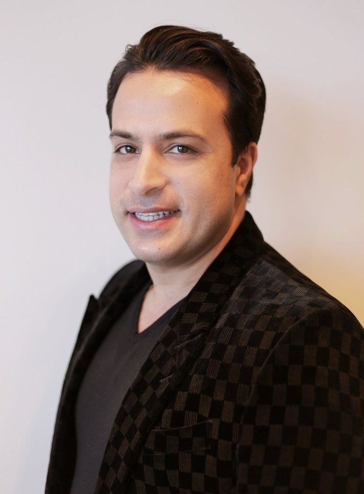 A man wearing a black checkered jacket and a black shirt smiles for the camera