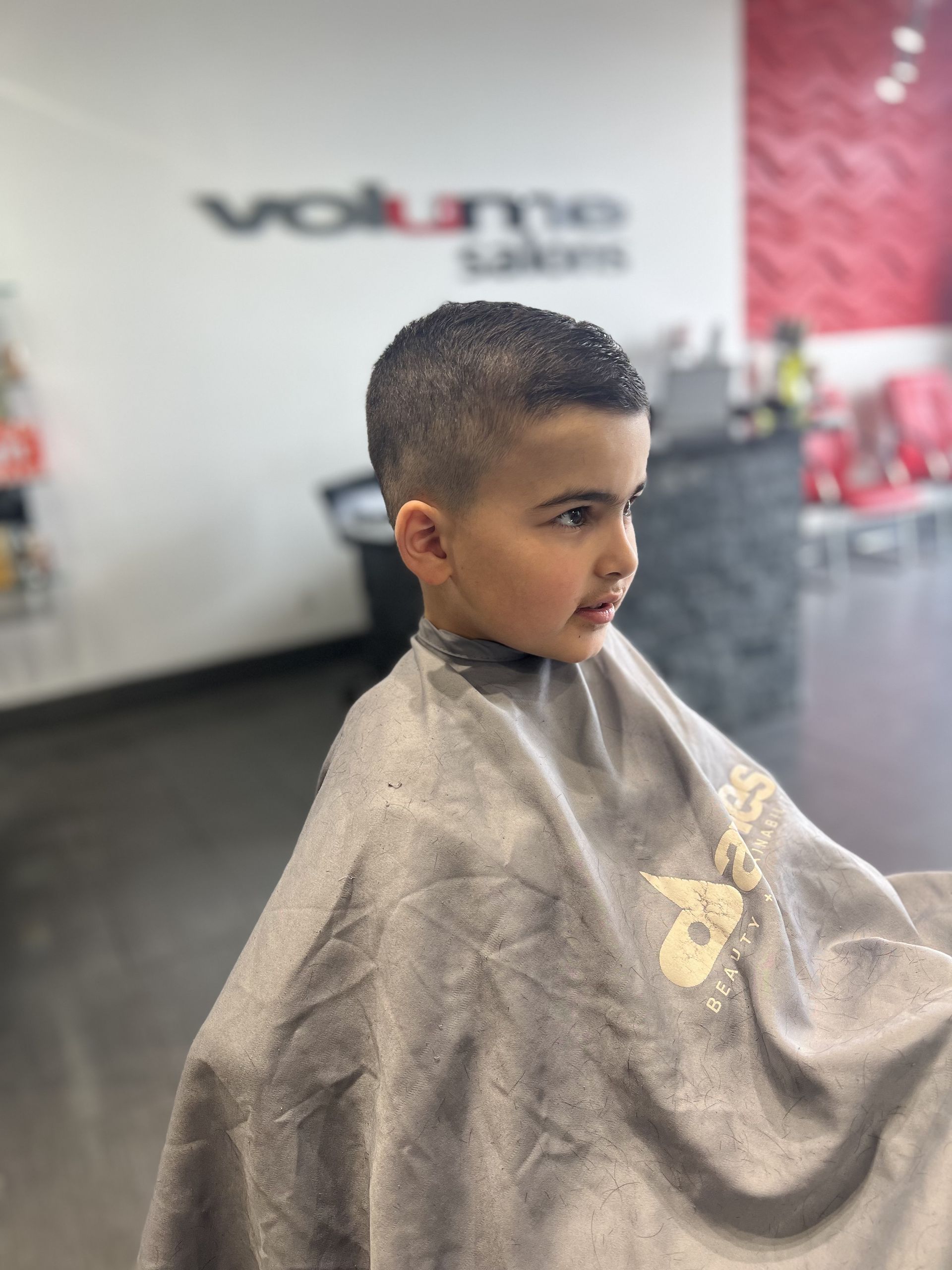 Young child with a fresh haircut at a hair salon, wearing a cape, looking to the side.