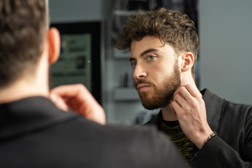 Man looking in a mirror, touching his beard. He is wearing a black blazer and has curly hair.