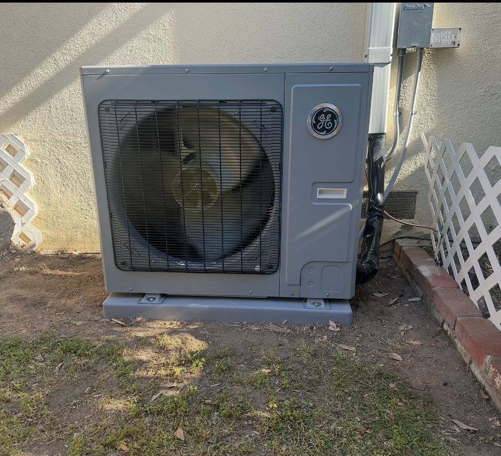 Gray GE heat pump unit, outdoors, on a concrete base, near a wall and grass.