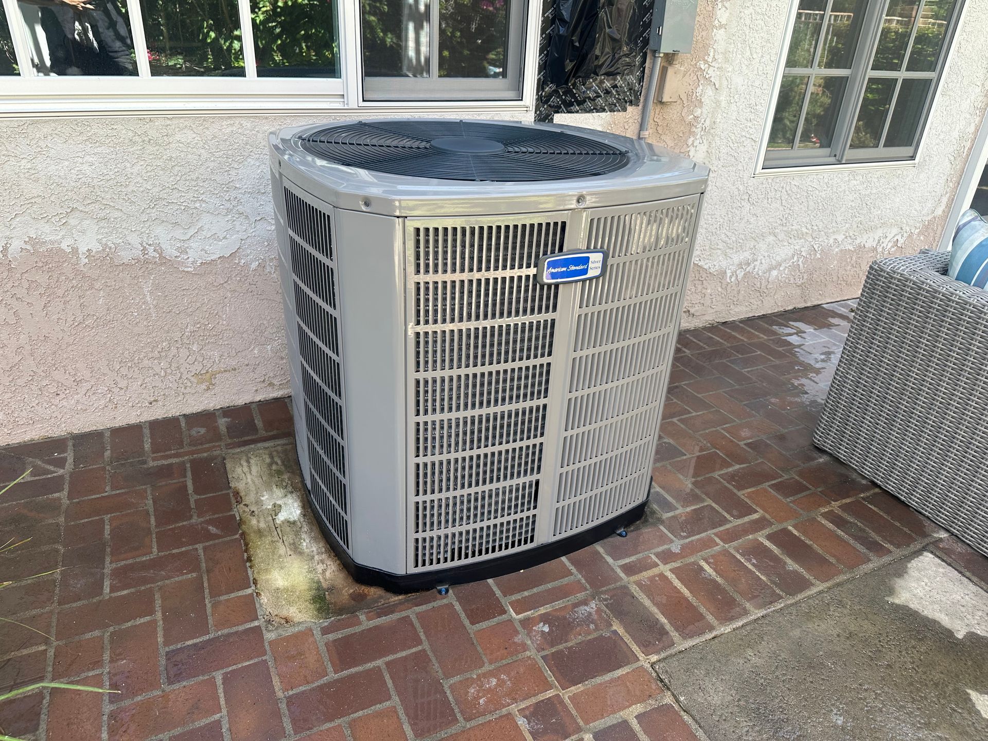 An outdoor air conditioning unit on a brick patio, near a window and outdoor furniture.