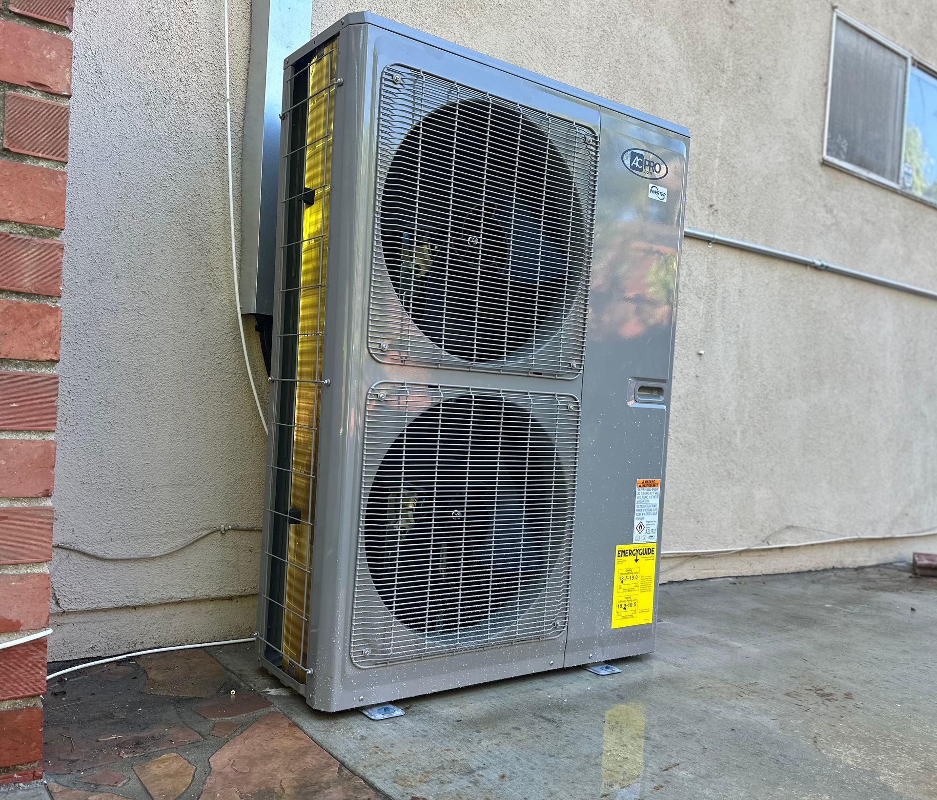 Gray air conditioning unit outside, mounted on concrete near a wall, with brick on the left.