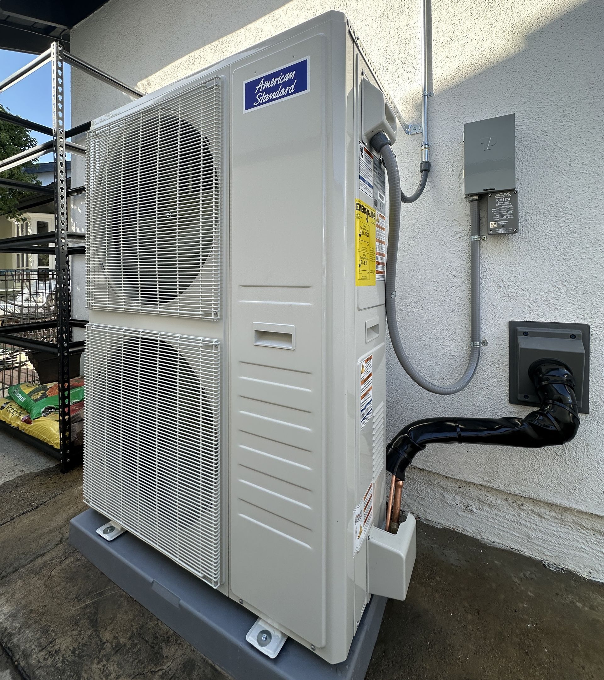 Exterior heat pump unit, beige, mounted on a concrete pad, against a stucco wall. Black vent and conduit visible.