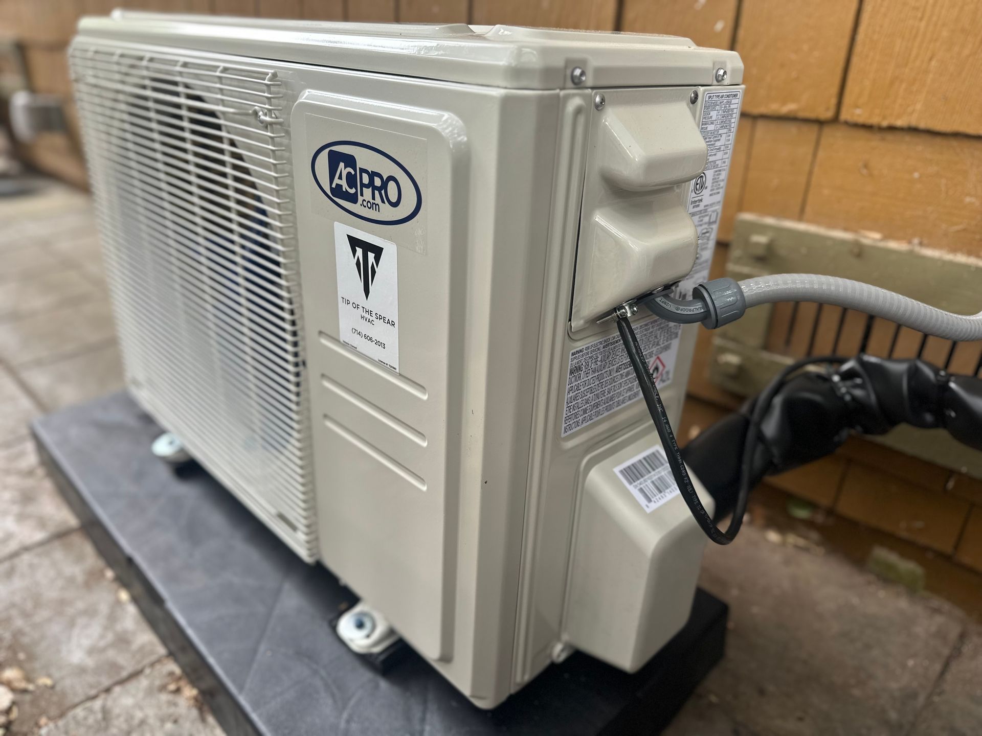 An outdoor heat pump unit, beige, mounted on a black pad against a brick wall.