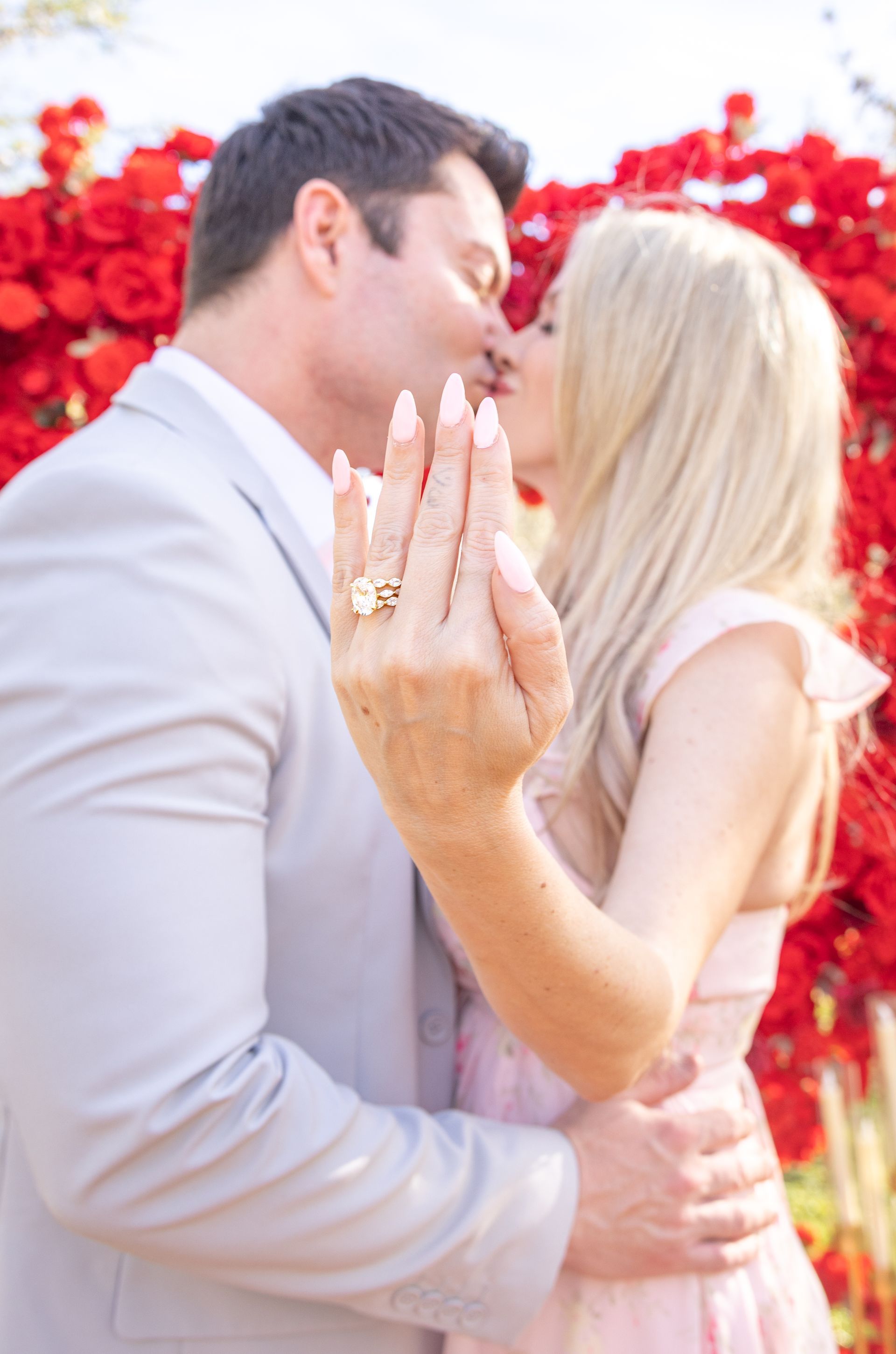 Two people pose for a wedding photo. One in a white dress and veil, the other in a green vest and bowtie, smiling in an outdoor setting.