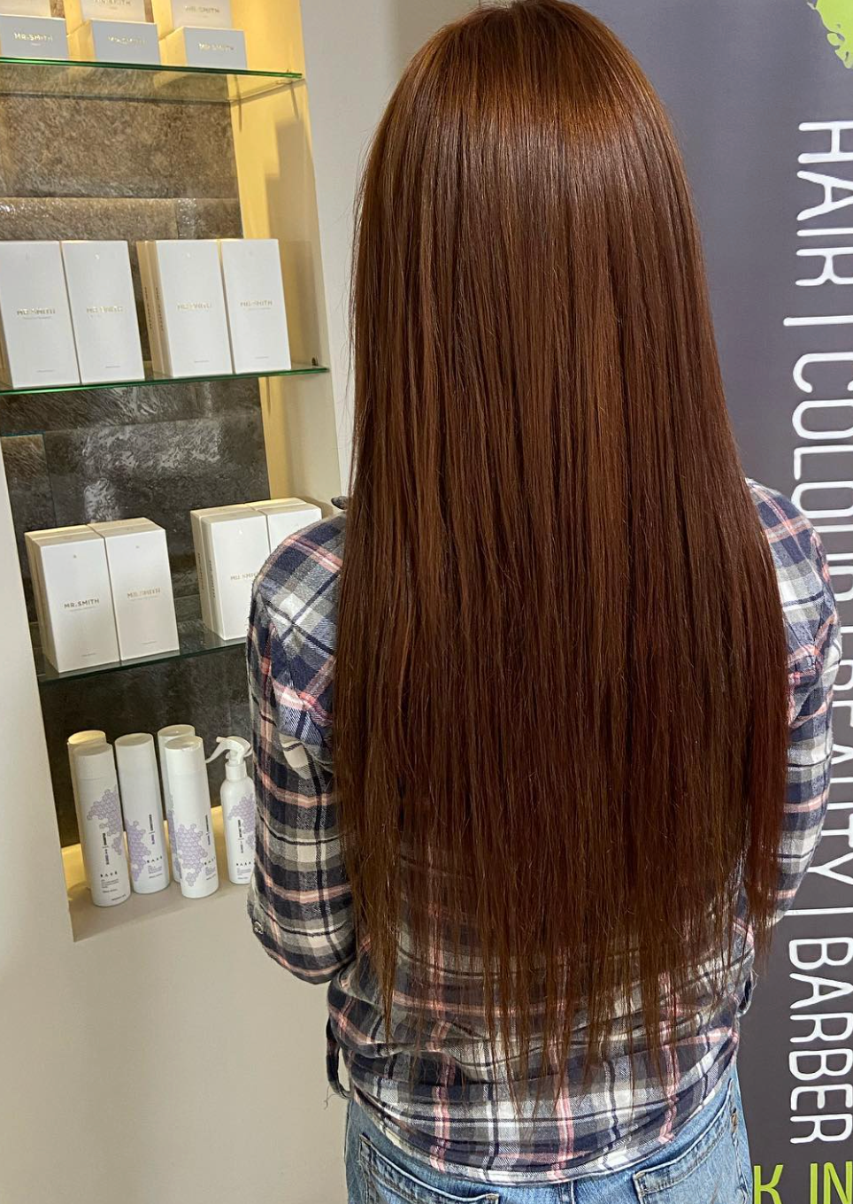 Woman with long, auburn hair stands in salon. She wears a plaid shirt and jeans. Products on shelf to her left.