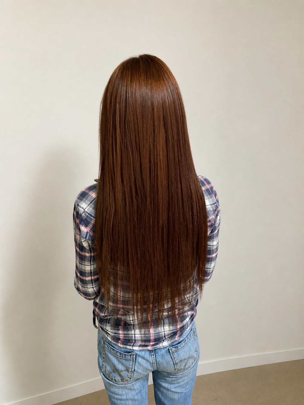 Person seen from behind with long straight brown hair, wearing a plaid shirt and jeans against a plain wall — Superb Hair in Wyong, NSW