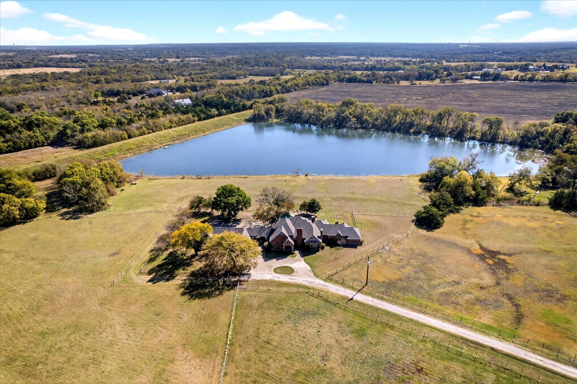 Aerial view: Large house with long driveway on grassy field near a lake. Trees surround the scene.