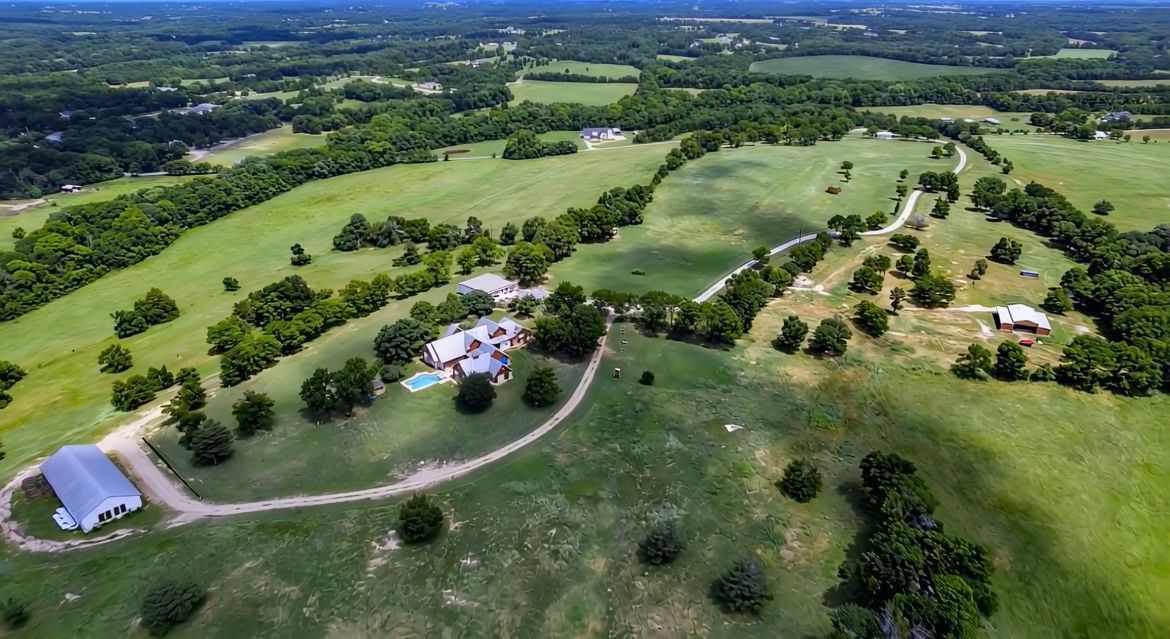 Aerial view of a large green property with buildings, trees, and fields on a sunny day.