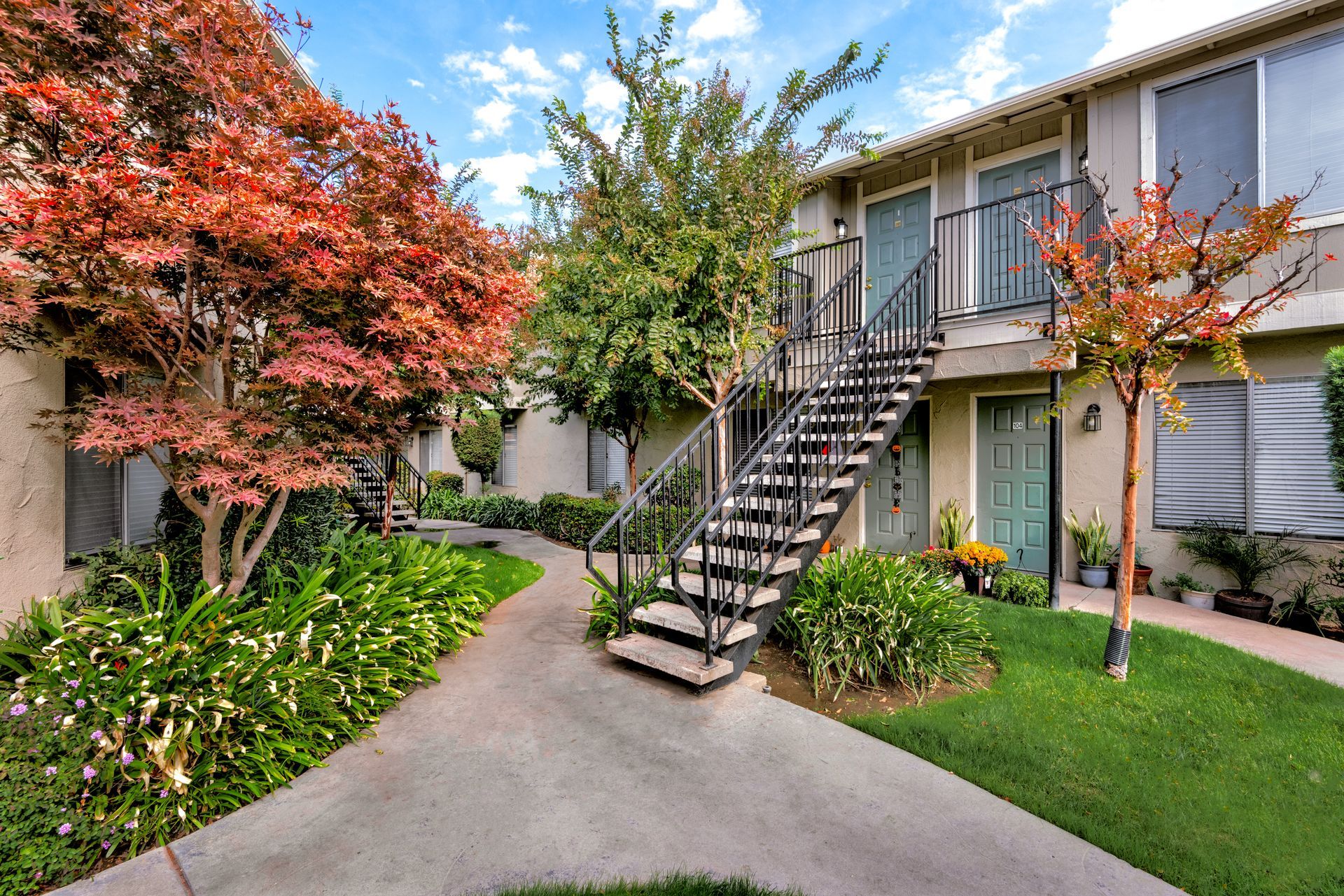 Apartment complex with stairs and walkway. Red and green trees surround the beige buildings.