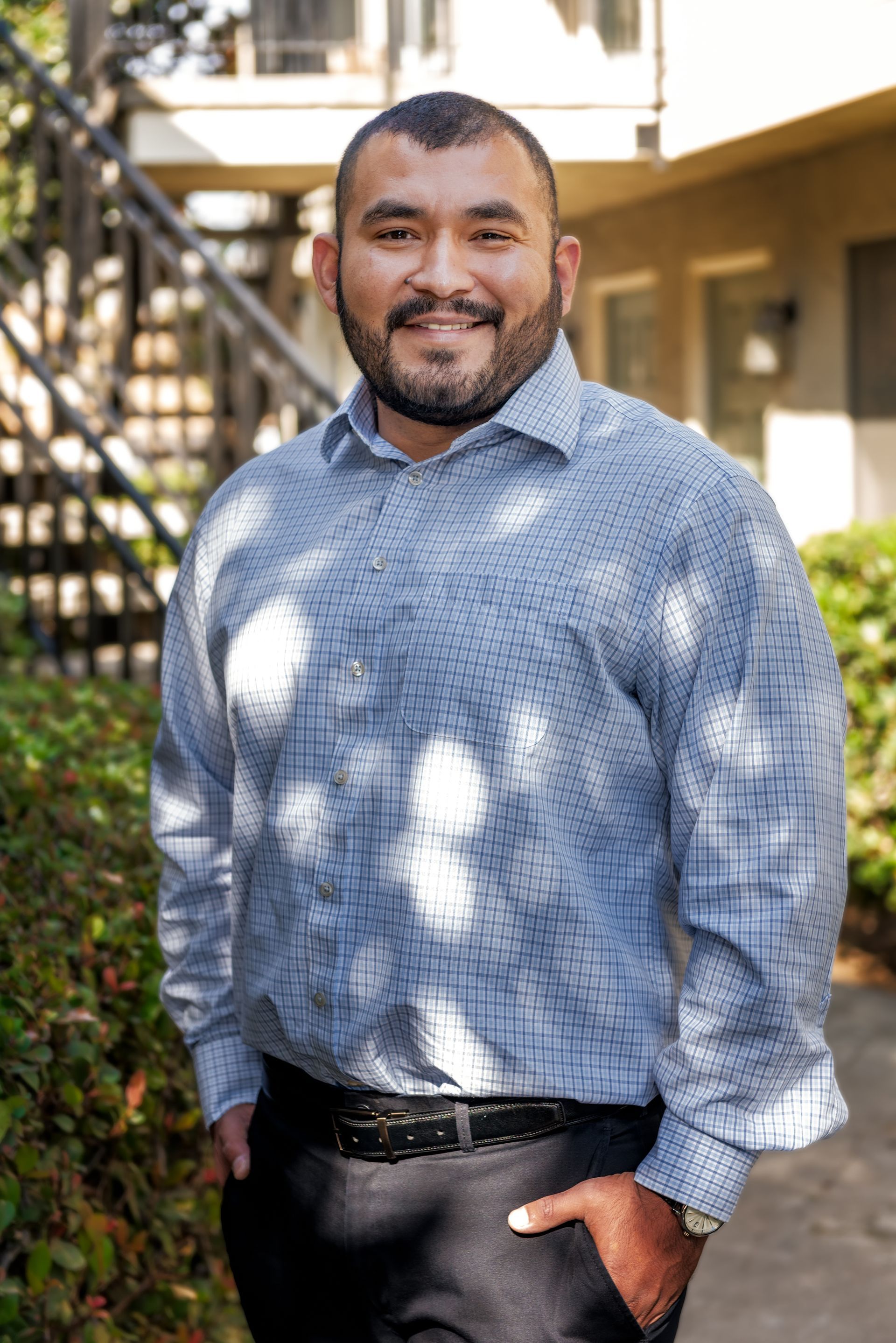 Man in blue button-up shirt, smiling, hands in pockets, standing outside near greenery and apartment building.