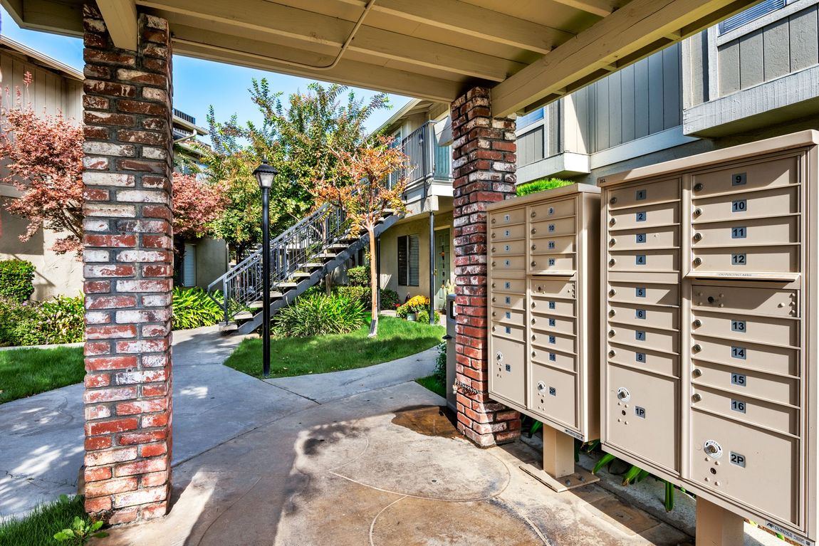 Mailboxes under a covered walkway with brick columns. Buildings and stairs in the background.