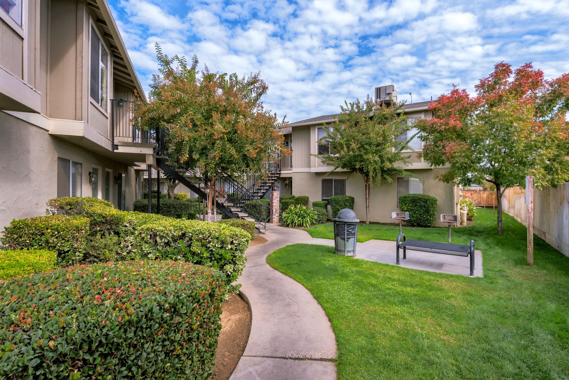 Pathway through a residential complex with trees, bushes, and benches on a sunny day.