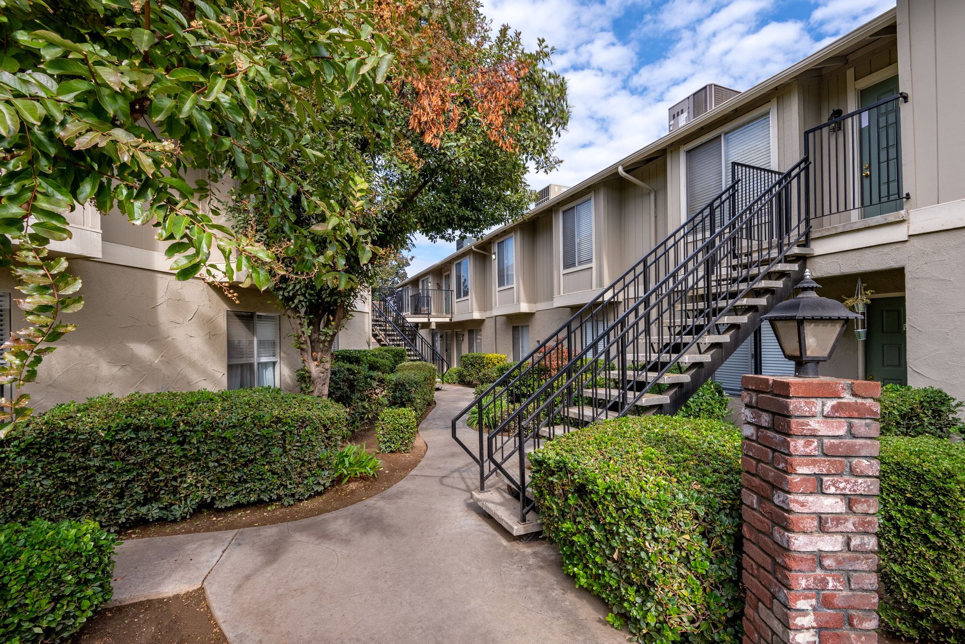 Apartment complex exterior with stairs, landscaping, and brick pillar under a blue sky with clouds.