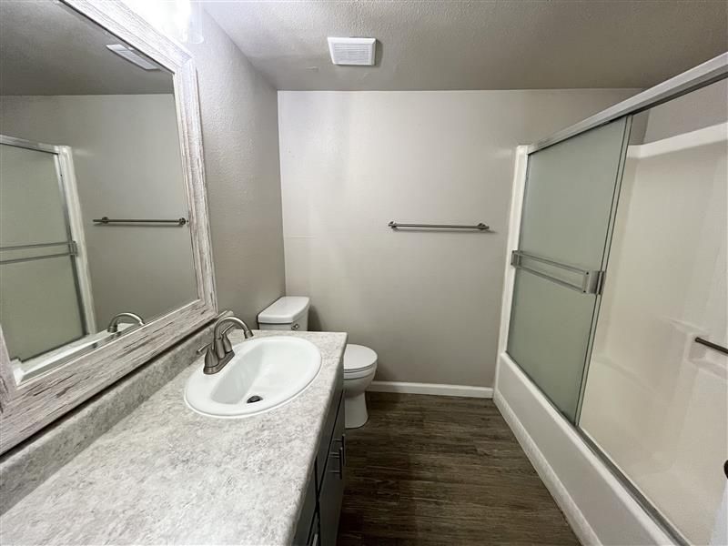 Bathroom with a vanity, toilet, bathtub, and shower. Features gray walls, a wood-look floor, and frosted shower doors.