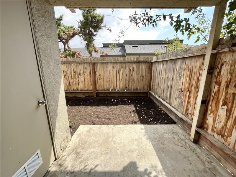 Concrete patio leads to a small yard enclosed by a weathered wooden fence. Brown soil, blue sky visible.