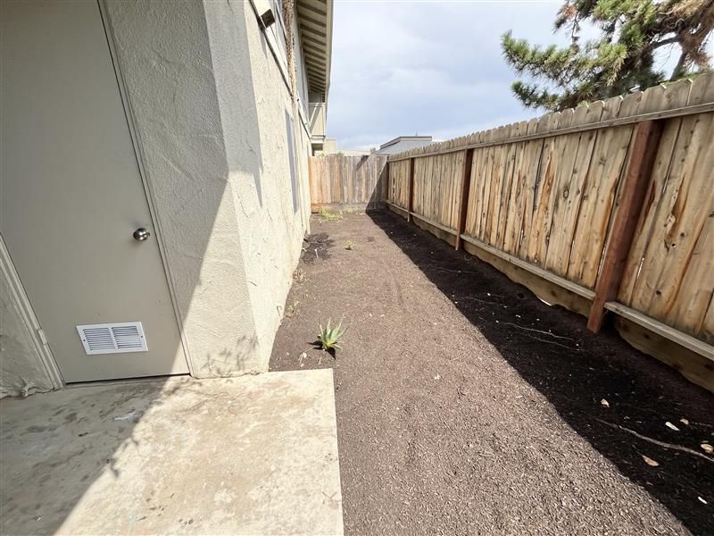 Narrow yard between a tan building and weathered wooden fence. Dark soil covers the ground.