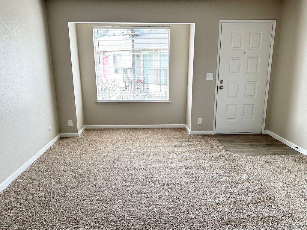 Empty room with beige walls, patterned carpet, window, and white door.
