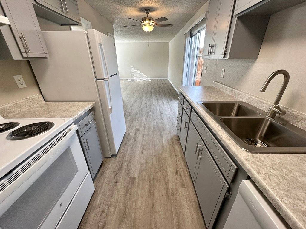 Kitchen with white appliances and cabinets, facing towards living area with light wood flooring.