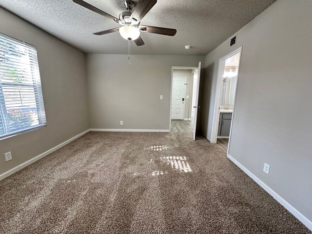 Bedroom with brown carpet, light gray walls, window, and ceiling fan. Doorway to bathroom.