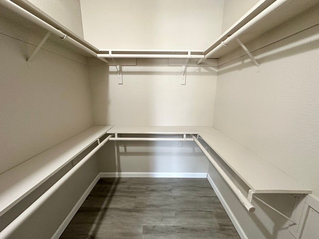 Empty walk-in closet with white shelves and rods against gray walls and wood-look floor.