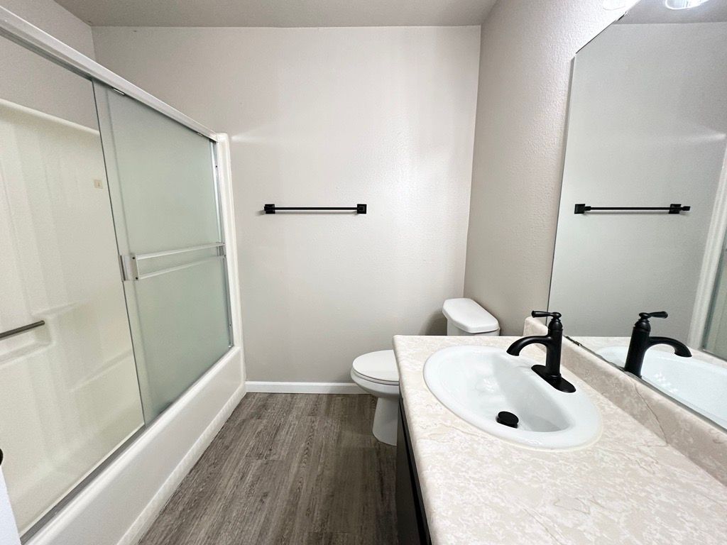Bathroom with white vanity, toilet, and tub. Black towel rack and faucet. Light gray walls and wood-look floor.