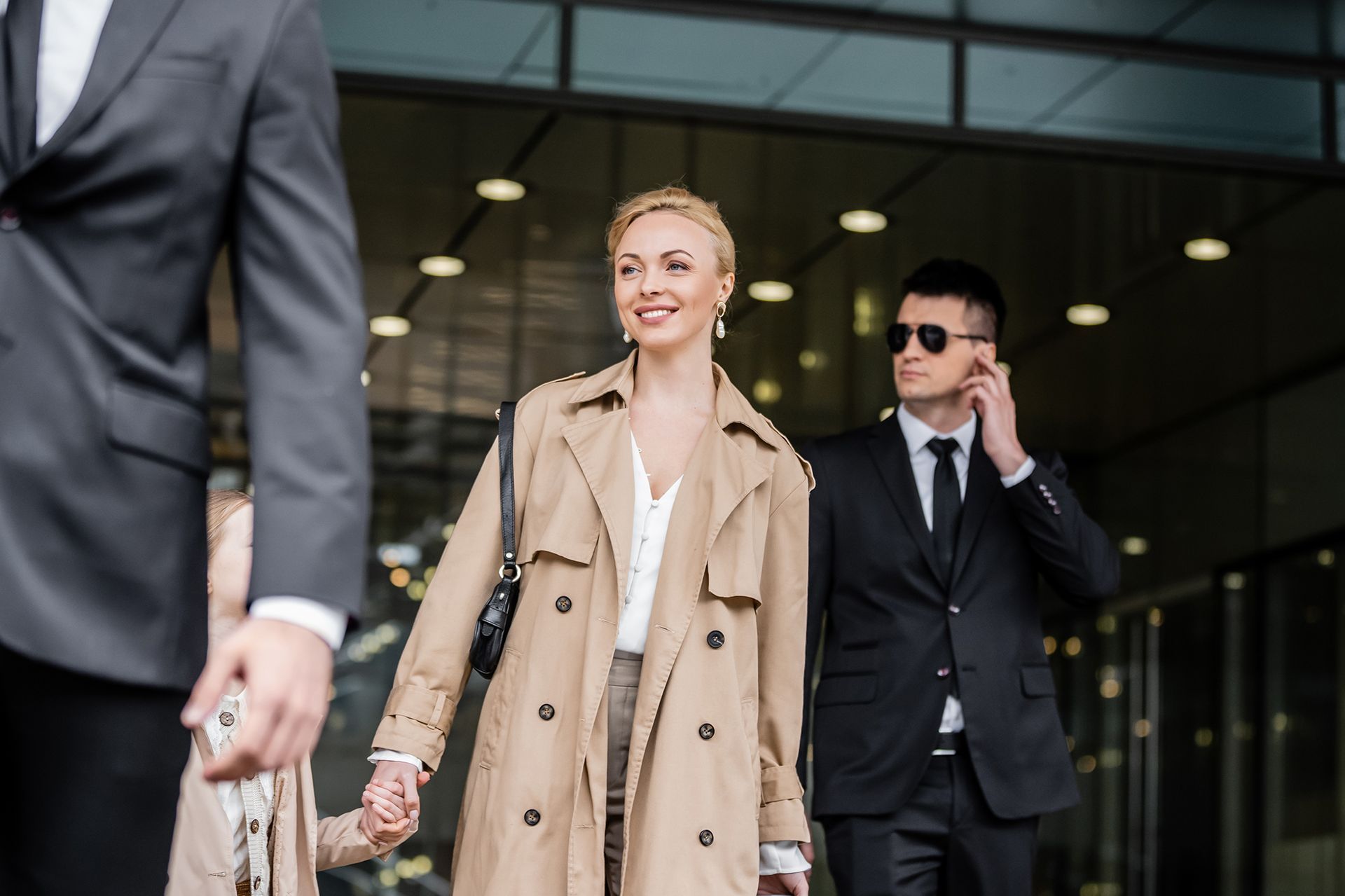 Woman in tan coat exits a building, flanked by two bodyguards in suits.