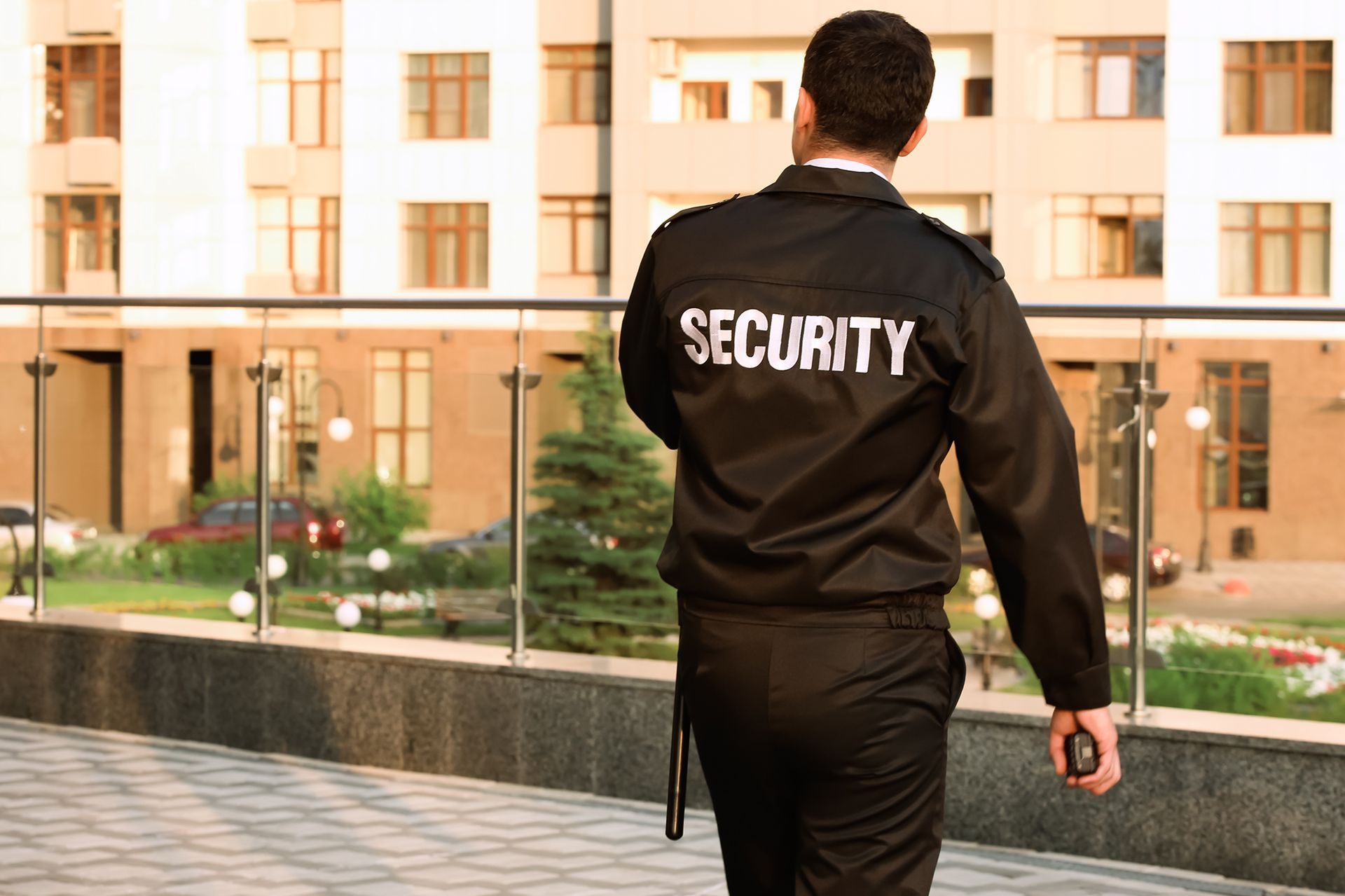 Security guard in black uniform walks, back to camera, outdoors by a building.