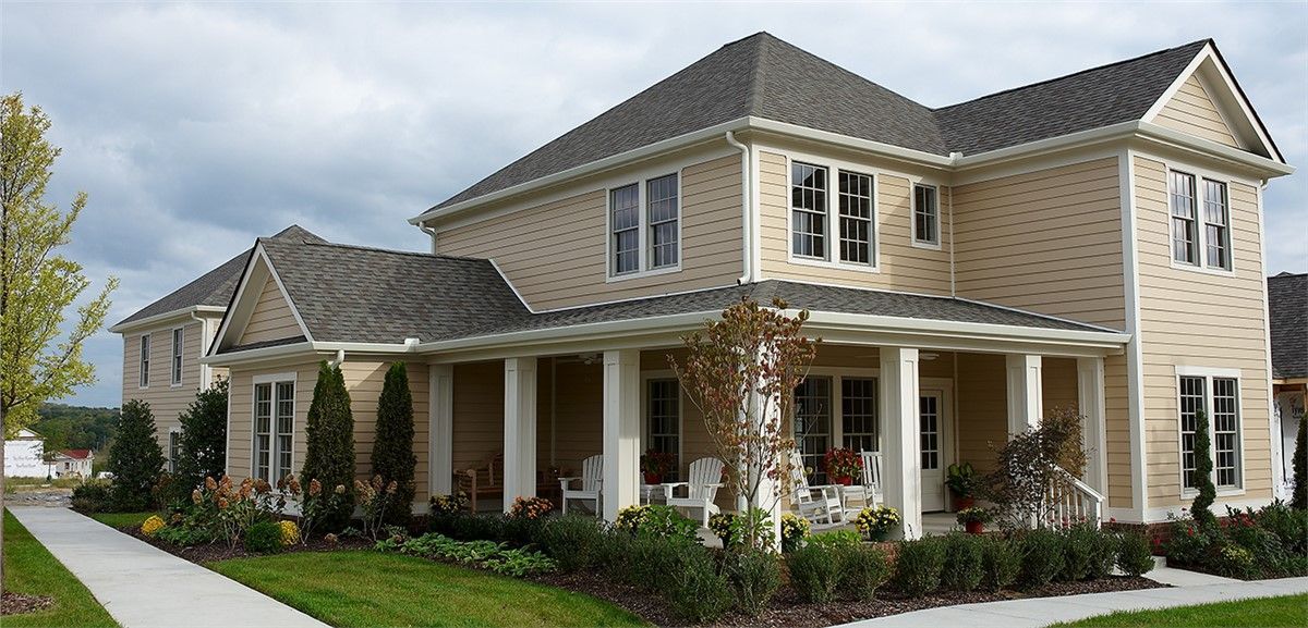 Beige two-story house with a porch and white columns, green lawn, and cloudy sky.